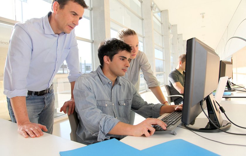 Erwachsenenschulung: Lehrer beugt sich am Computer von hinten über einen seiner Schüler und erklärt ihm etwas