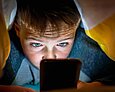 Ein etwa zehnjähriger Junge liegt unter einer Bettdecke und schaut erstaunt bis verstört in sein Handy.