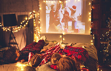 Eine Familie liegt auf dem Sofa und schaut auf der Leinwand einen Film. Um sie herum viele Lichterketten und Weihnachtsdekoration.