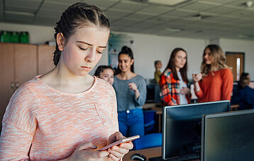 Cybermobbing: Mädchen schaut traurig auf ihr Handy. Im Hintergrund lästern und lachen Schülerinnen über sie.
