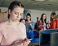 Cybermobbing: Mädchen schaut traurig auf ihr Handy. Im Hintergrund lästern und lachen Schülerinnen über sie.