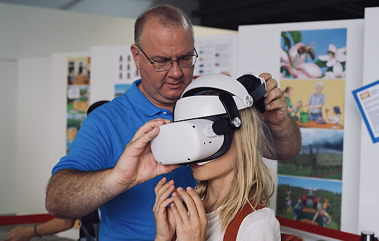 Älterer Mann mit Brille und blauen Hemd setzt blonden Mädchen mit weißem Hemd eine VR-Brille auf.