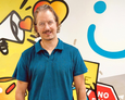 Christian Heneka Ein Mann mittleren Alters mit dunkelblauem Polohemd steht vor einer hellgrauen Wand mit Graffiti-Logos des Projekts Bitte Was?!