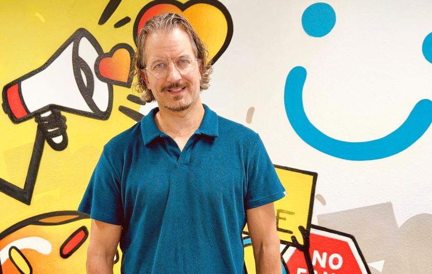 Ein Mann mittleren Alters mit dunkelblauem Polohemd steht vor einer hellgrauen Wand mit Graffiti-Logos des Projekts Bitte Was?!