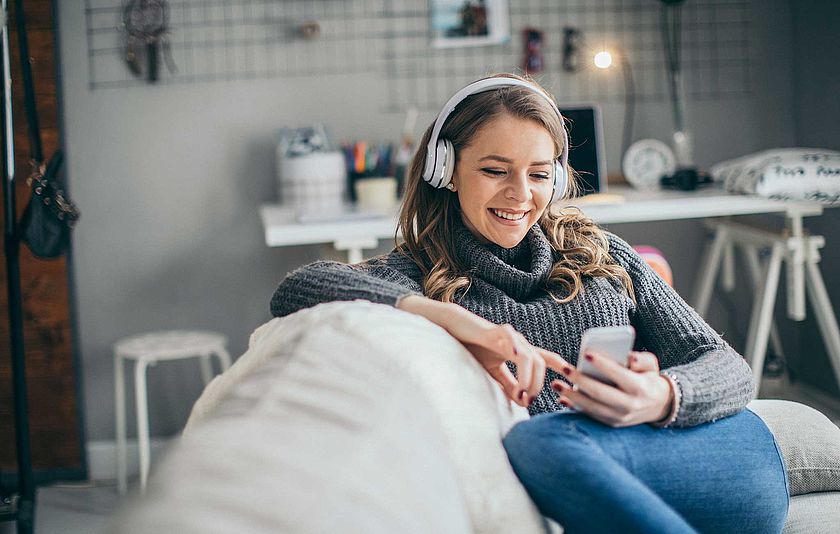 Junge Frau sitzt mit Kopfhörer und Smartphone auf dem Sofa.