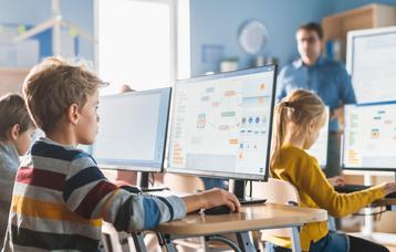 Mehrere Grundschüler arbeiten hoch konzentriert an ihrem PC. Auf dem Bildschirm sind Flußdiagramme zu sehen. Im Hintergrund steht der Lehrer und beobachtet seine Klasse.