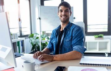 Lächelnder, sympathischer Softwareentwickler, der während der Arbeit am Computer in einem modernen Start-up-Büro in die Kamera blickt.