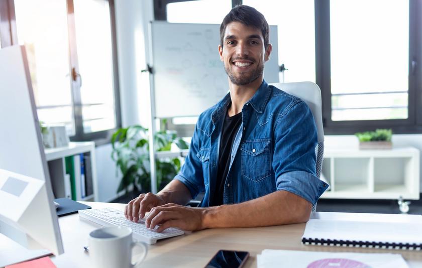 Lächelnder, sympathischer Softwareentwickler, der während der Arbeit am Computer in einem modernen Start-up-Büro in die Kamera blickt.