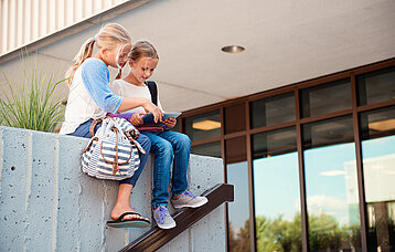 Grundschülerinnen mit Tablet sitzend auf einer Mauer