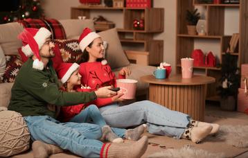 Ein Mann, ein Mädchen und eine Frau mit Weihnachtsmützen auf dem Kopf sitzen im Wohnzimmer und halten eine Fernbedienung und Popcorn in der Hand.