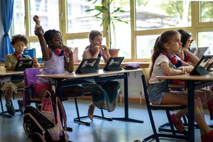 Mehrere Schülerinnen und Schüler in einer Grundschule sitzen an ihren Tischen und ein Mädchen streckt.