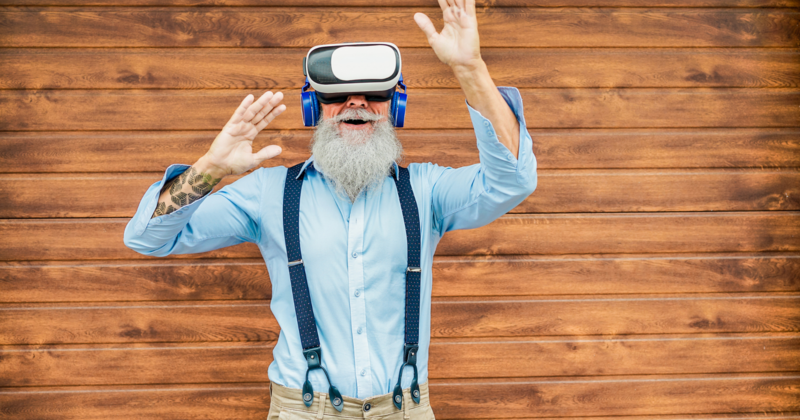 Ein Senior mit grauen Haaren und grauem Bart hat eine Virtual Reality Brille und blaue Kopfhörer an und hat Spaß