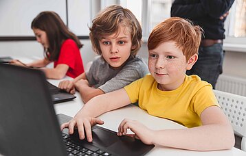 Zwei Grundschüler sitzen hoch konzentriert und motiviert vor einem Laptop. Einer der Jungen hat feuerrotes Haar und Sommersprossen.