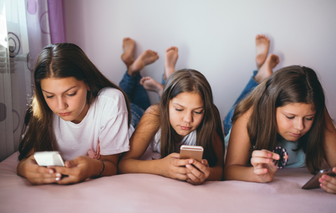 Drei junge Mädchen liegen nebeneinander bäuchlings auf einem Bett und schauen auf ihre Handys in ihren Händen. 