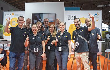 Zehn Frauen und Männer in schwarzen Polo-T-Shirts mit dem Logo des Landesmedienzentrums zeigen lachend "Daumen hoch" in die Kamera. Im Hintergrund ist ein weiß-orange-farbener Messestand zu sehen.