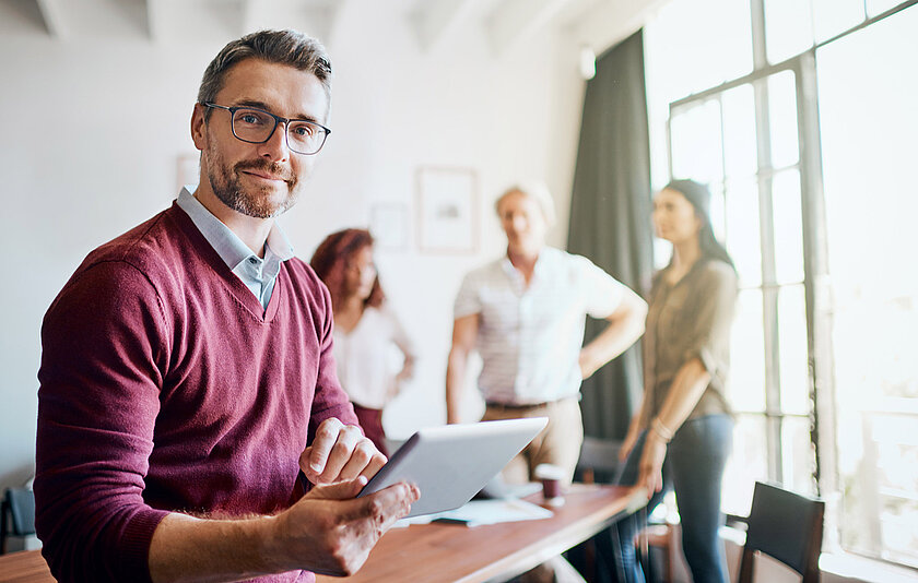 Lehrer mit iPad, im Hintergrund verschwommen Kolleginnen und Kollegen im Lehrerzimmer