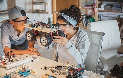 Zwei Teenager (Junge und Mädchen) arbeiten in der Werkstatt an einem Roboter-Projekt.