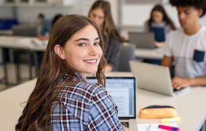 Lächelnde Schülerin vor ihrem PC in die Kamera lächelnd. Im Hintergrund sind andere Schüler/-innen zu sehen.