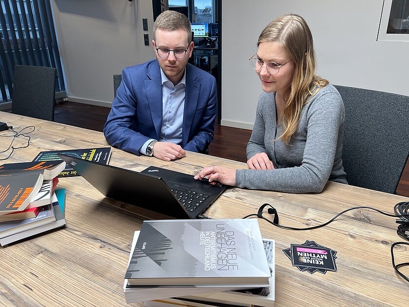 Ronja Schonscheck im Interview über die Plattform OY VEY gegen antisemitische Verschwörungsmythen Ein junger Mann im blauen Anzug und eine junge Frau in einer grauen Weste sitzen an einem Holztisch und blicken auf einen Laptop, der vor ihnen steht. Im Vordergrund liegen Bücher.