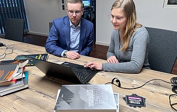 Ein junger Mann im blauen Anzug und eine junge Frau in einer grauen Weste sitzen an einem Holztisch und blicken auf einen Laptop, der vor ihnen steht. Im Vordergrund liegen Bücher.