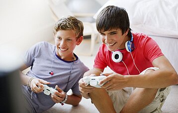 Zwei Teenager-Jungs sitzen auf dem Boden und halten Controller für eine Computerspielkonsole in der Hand. 