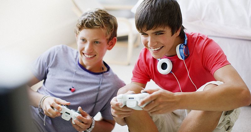 Zwei Teenager-Jungs sitzen auf dem Boden und halten Controller für eine Computerspielkonsole in der Hand. 