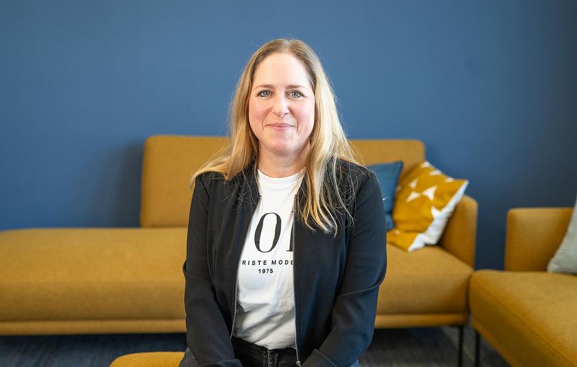 Junge Frau mit blonden langen Haaren sitzt vor gelbem Sofa und blauer Wand.