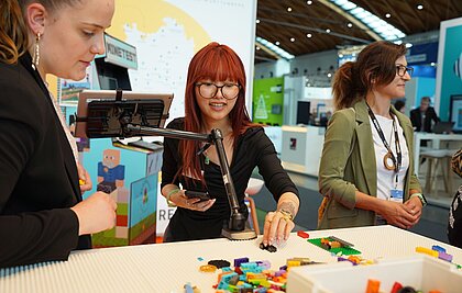 Eine junge Frau mit roten Haaren baut auf einem Tisch mit einer Legoplatte mit Lego-Bausteinen. Daneben steht eine andere Frau und überlegt. Zwischen Ihnen ist ein Tablet an einer Halterung befestigt.