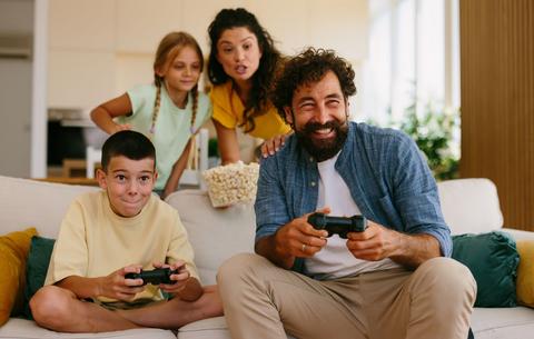 Eine vierköpfige Familie sitz mit Gamecontrollern auf einem Sofa und spielt gemeinsam.