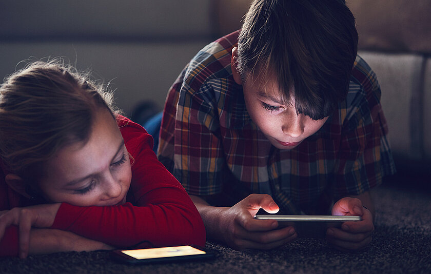 Ein Junge liegt auf dem Bauch und schaut übermüdet in ein Tablet, während die Schwester daneben auf dem Boden schläft.