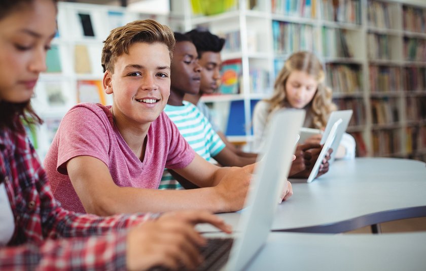 Jugendlicher sitzt mit anderen Schülerinnen und Schülern in einer Bibliothek und lächelt in die Kamera.
