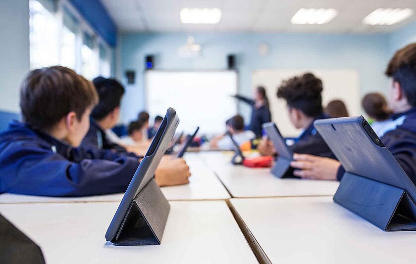 Blick von hinten im Klassenzimmer nach vorne ans Whiteboard. Alle Schüler haben ihr Tablet aufgestellt und schauen an die Tafel.