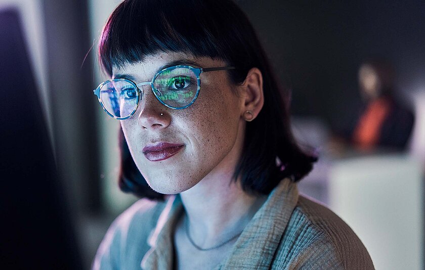 Junge Frau mit Brille lächelt in den Bildschirm. Der Hintergrund ist dunkel.