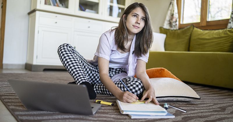 Teenagerin sitzt mit ihrem Laptop auf dem Boden und schreibt auf einen Block.