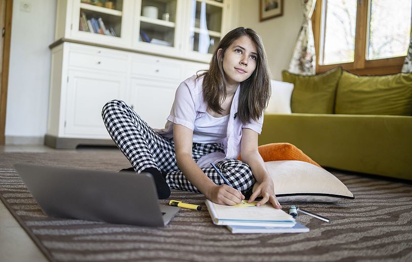 Teenagerin sitzt mit ihrem Laptop auf dem Boden und schreibt auf einen Block.