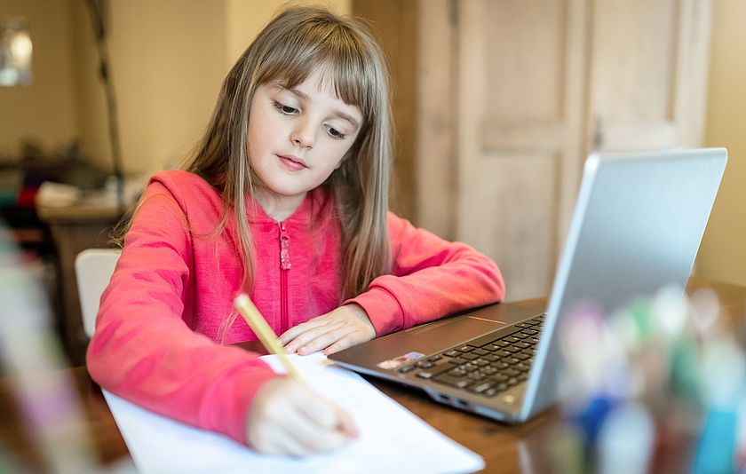 Neben einem Laptop schreibt eine Grundschülerin auf ein Blatt Papier