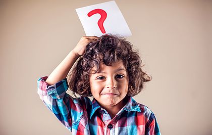 Ein Junge mit Locken hält ein Papier auf dem ein Fragezeichen steht hoch und lacht in die Kamera