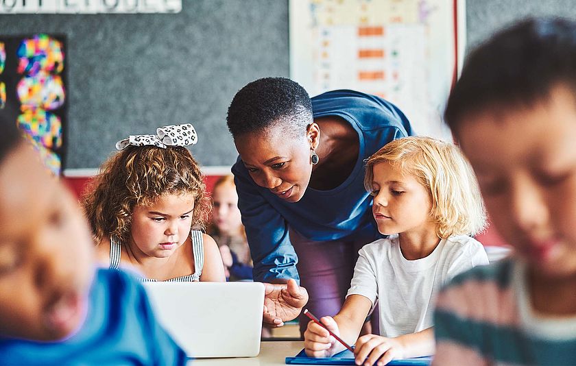 Man sieht eine weibliche Lehrkraft, die zwei Schülerinnen im Unterricht etwas auf dem Tablet zeigt.