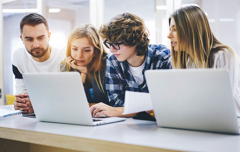 Vier Journalistinnen und Journalisten sitzen vor zwei Laptops und stecken nachdenklich die Köpfe zusammen.