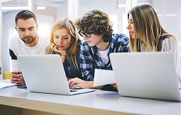 Vier Journalistinnen und Journalisten sitzen vor zwei Laptops und stecken nachdenklich die Köpfe zusammen.