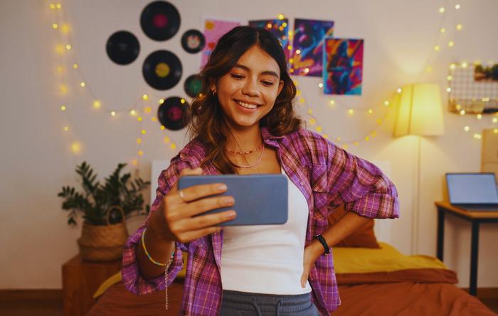 Alterskontrolle per KI auf Meta-Plattformen Teenager-Mädchen posiert in ihrem Jugendzimmer vor einem Smartphone.