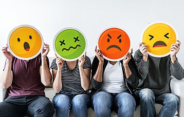 Personen halten sich negative Emojis vor ihre Gesichter