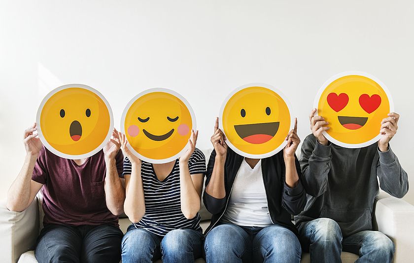 Vier Personen halten unterschiedliche Emojis aus Pappe vor ihr Gesicht.