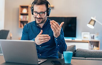 Junger Mann mit Kopfhörern gestikuliert vor dem Laptop bei einer Videokonferenz.