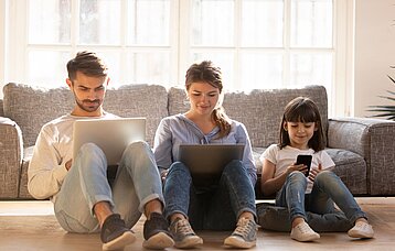 Vater, Mutter und Kind sitzen mit elektronischen Geräten in der Hand nebeneinander auf dem Sofa.