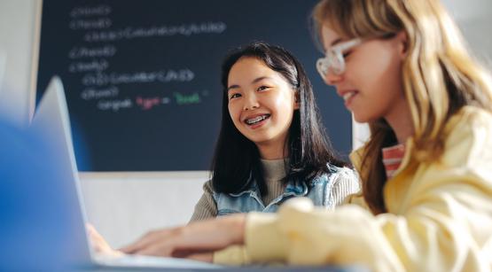Ein Eine Schülerin mit Zahnspange und braunen Haaren lacht in die Kamera. Neben ihr sitzt ihre Mitschuelerin. Die beiden recherchieren etwas mit dem Laptop.