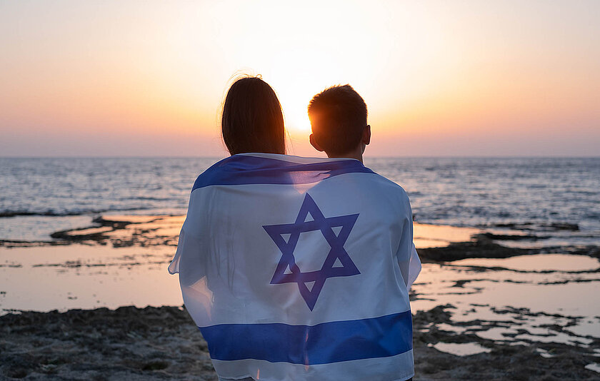 Teenager, junge Frauen und Männer mit der Flagge Israels über den Schultern beim Sonnenuntergang über dem Meer in Israel. 