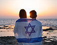 Teenager, junge Frauen und Männer mit der Flagge Israels über den Schultern beim Sonnenuntergang über dem Meer in Israel. 
