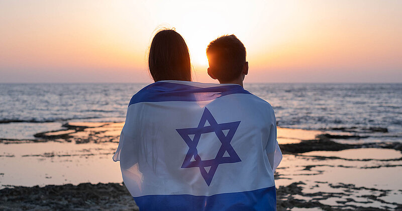 Teenager, junge Frauen und Männer mit der Flagge Israels über den Schultern beim Sonnenuntergang über dem Meer in Israel. 