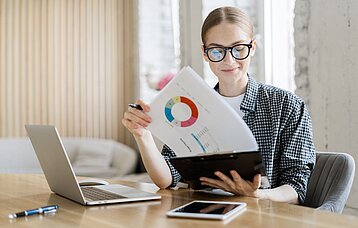 Junge Frau mit schwarzer Brille sitzt an einem Schreibtisch. Sie hält ein Klemmbrett mit Statistiken in der Hand. Vor ihr stehen ein Laptop und ein Tablet.